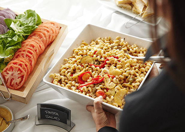 Person scooping pasta salad from a corporate event catering spread